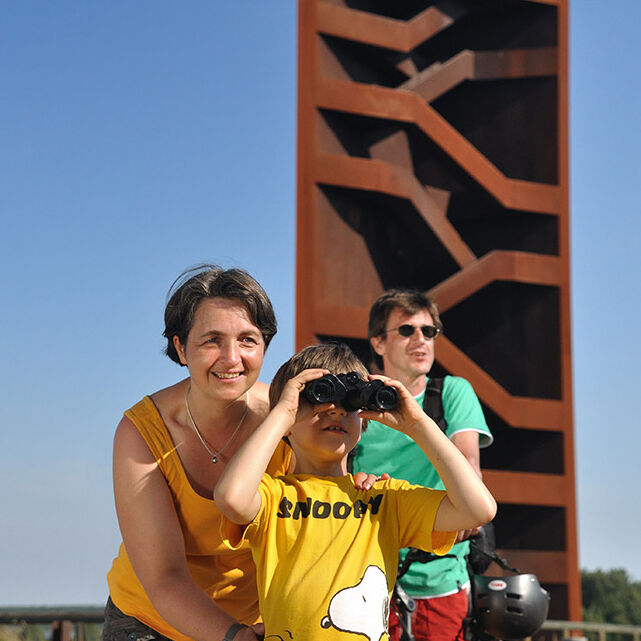 Eine Familie mit Kind erkundet die alten Industriebrachen in der Lausitz. Im Hintergrund befindet sich ein künstlerischer Turm vor blauem Himmel.