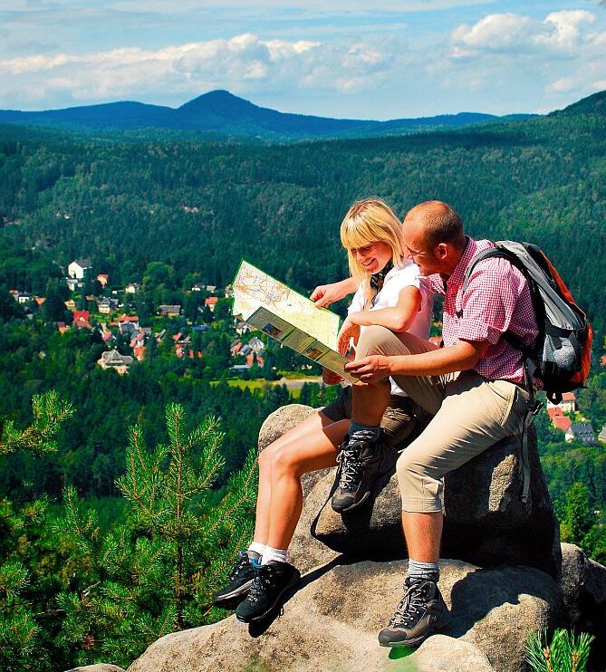 Zwei Personen stehen an Gipfel und lesen eine Wanderkarte. Im Hintergrund eine Gebirgslandschaft.