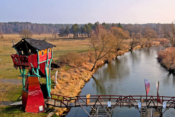 Fluss mit langer Holzbrücke und einer Art Baumhaus am Brückenende, Vogelperspektive 